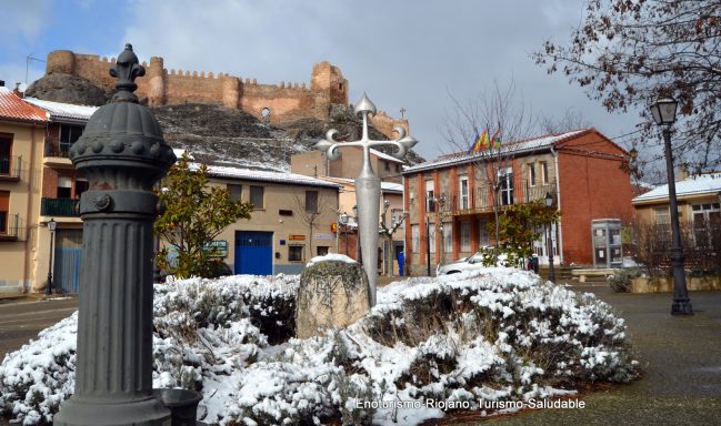 Plaza con nieve, monumento en el centro y castillo al fondo. Edificios coloridos alrededor.